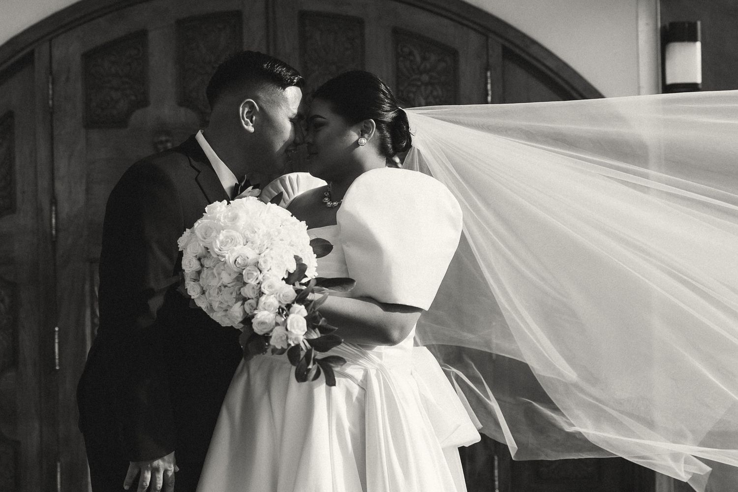 Black and white photo of a bride and groom sharing a kiss, her veil flowing in the wind, holding a white bouquet.