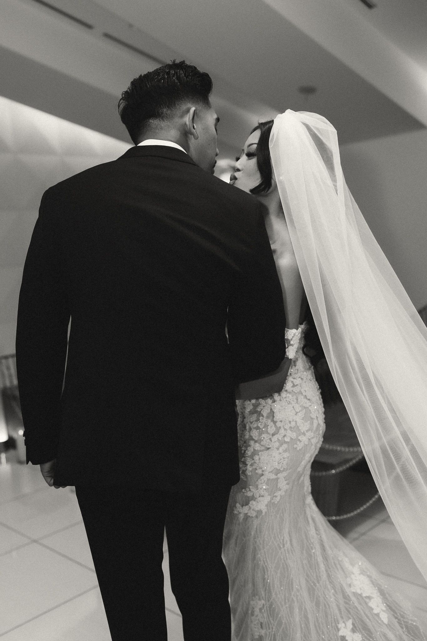 Black and white photo of a bride and groom sharing an intimate moment, bride in lace gown with veil, groom in tuxedo.