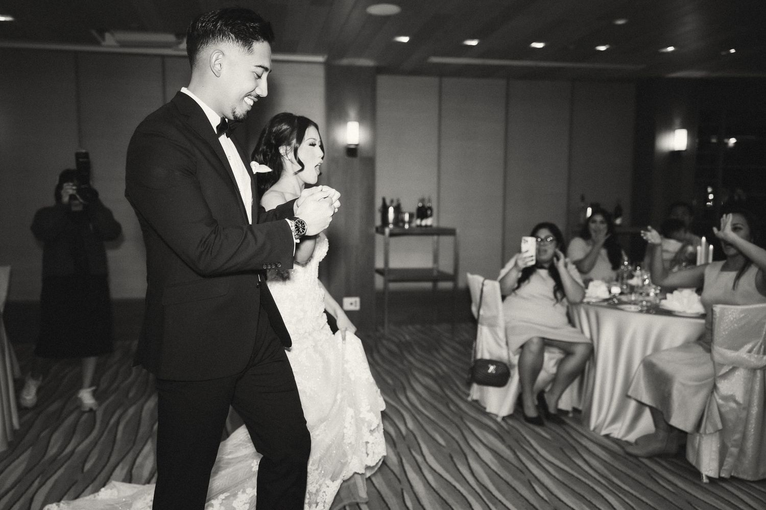 Black and white photo of a couple sharing their first dance at a wedding reception with guests watching and photographing.