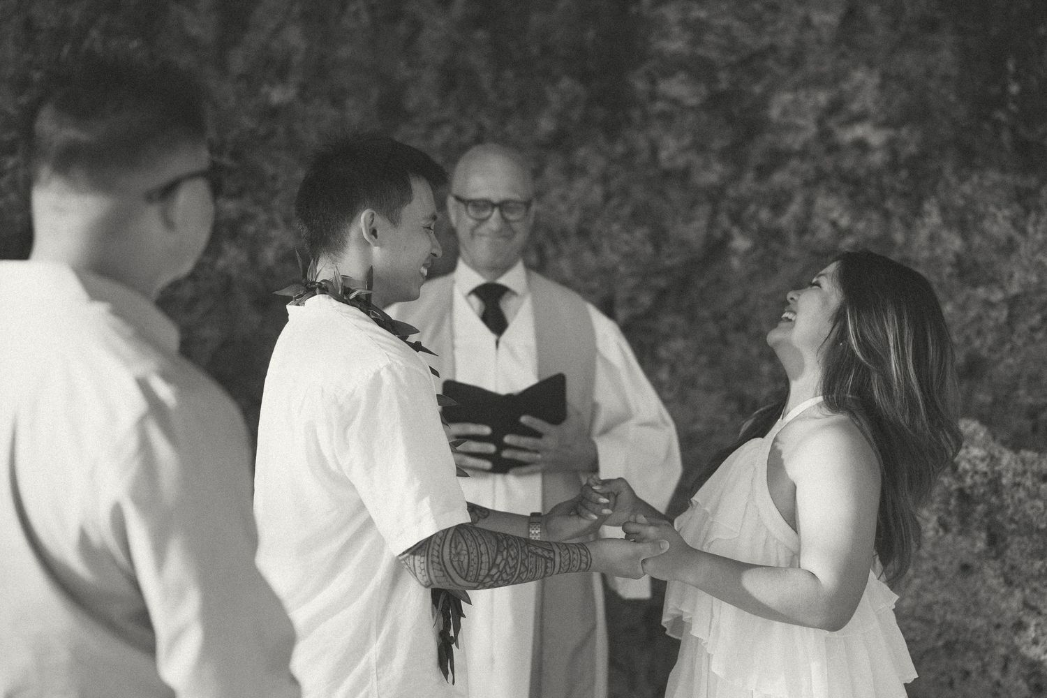 Black and white wedding ceremony photo with couple exchanging vows before officiant outdoors.