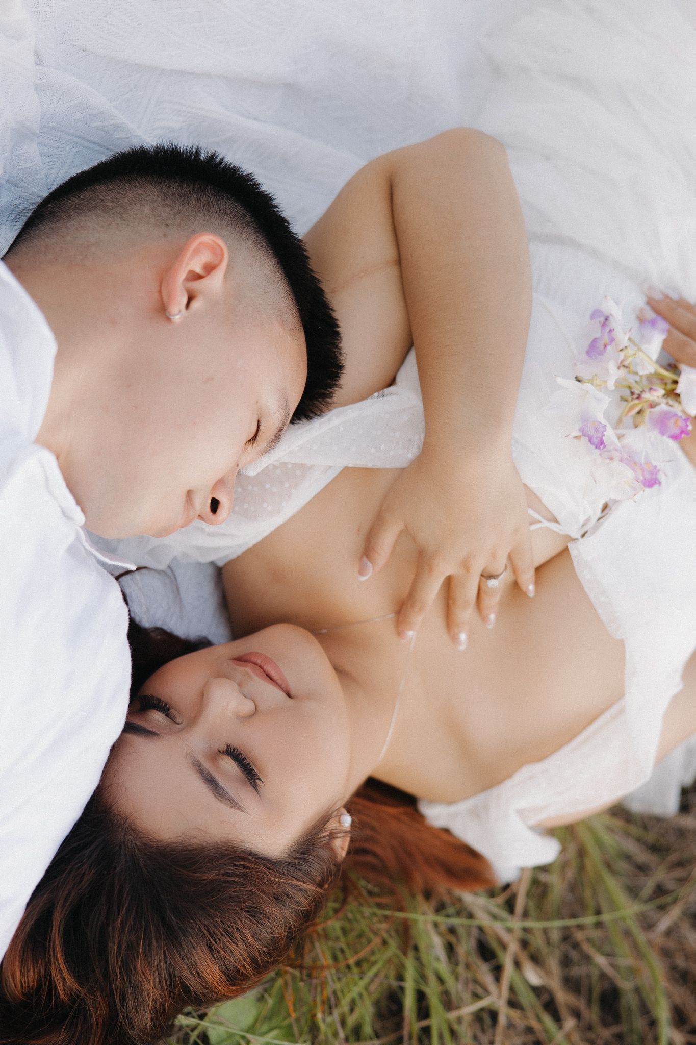 Couple lying in grass, man nuzzling woman's neck, romantic outdoor portrait with white fabric and purple flowers.