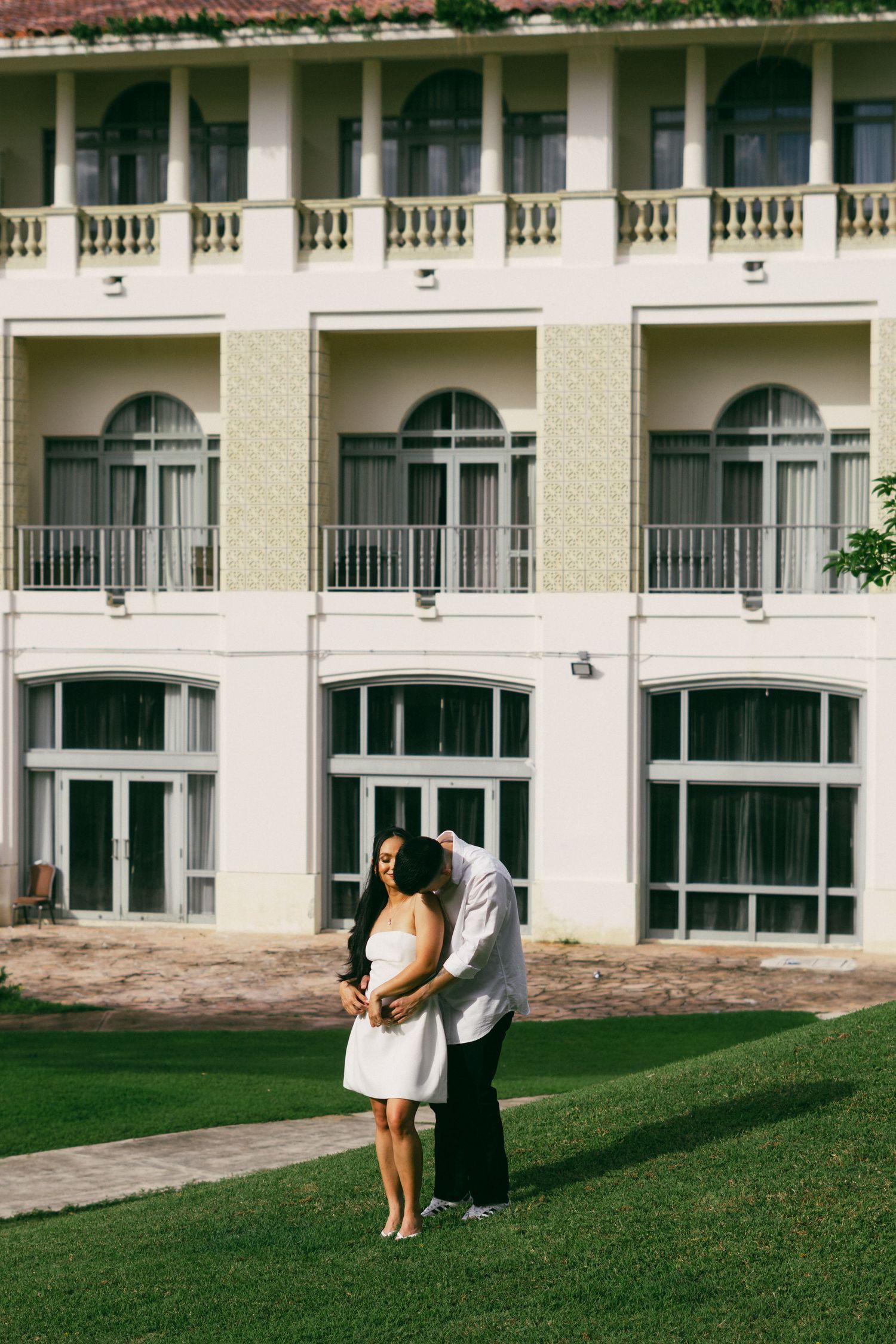 Couple embracing on green lawn in front of elegant white colonial building with arched windows and balconies.