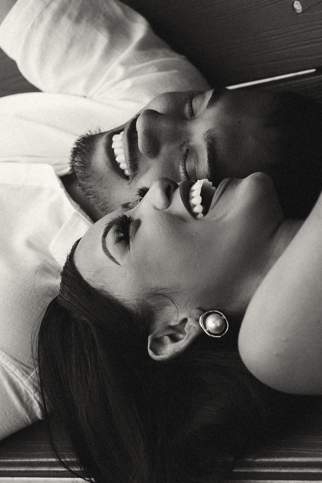 Black and white photo of a happy couple lying down, laughing and smiling together in an intimate romantic moment.