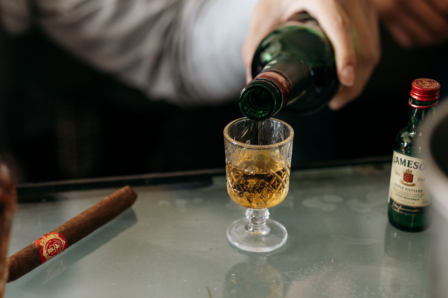 Whiskey being poured from a green bottle into a crystal glass beside a cigar and Jameson miniature bottle.