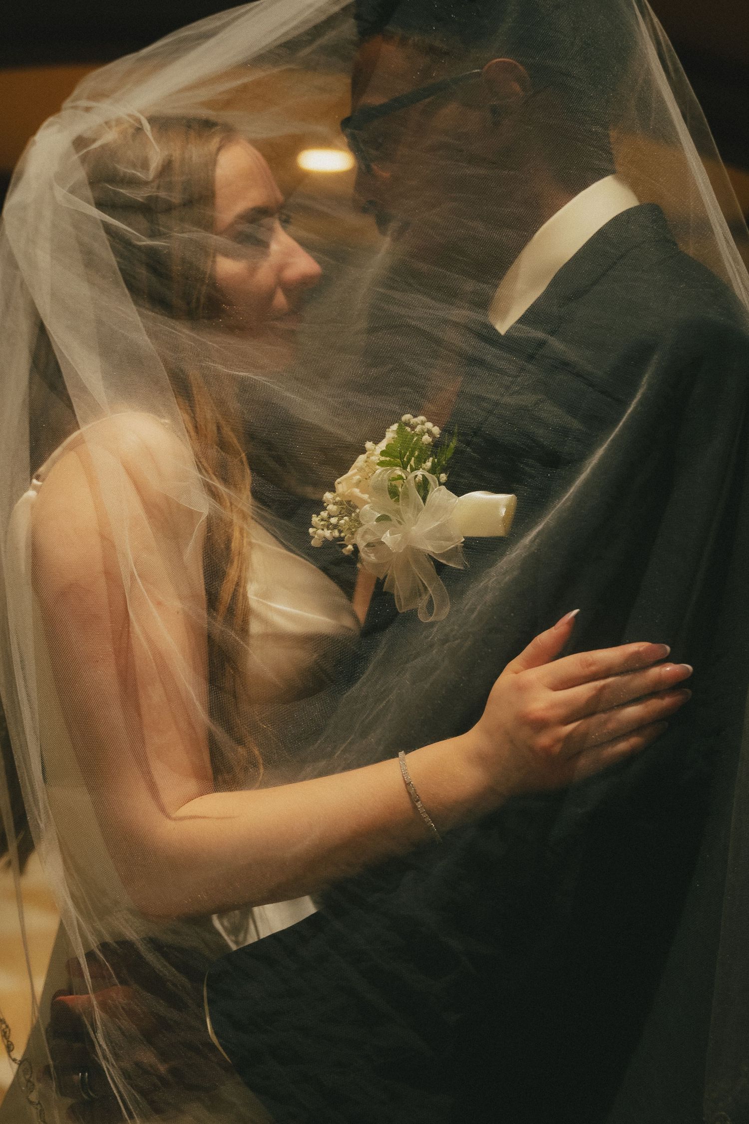Bride and groom share an intimate moment under a flowing veil, with a white boutonniere visible.