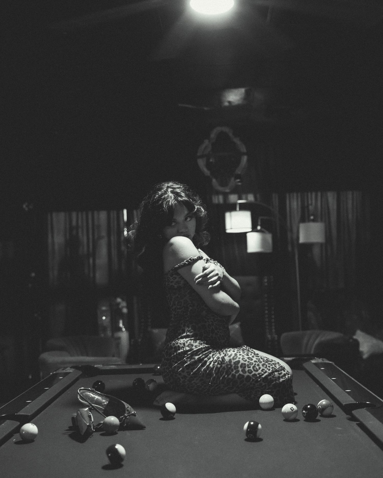 Black and white photo of a woman sitting on a pool table surrounded by billiard balls in a dimly lit room.