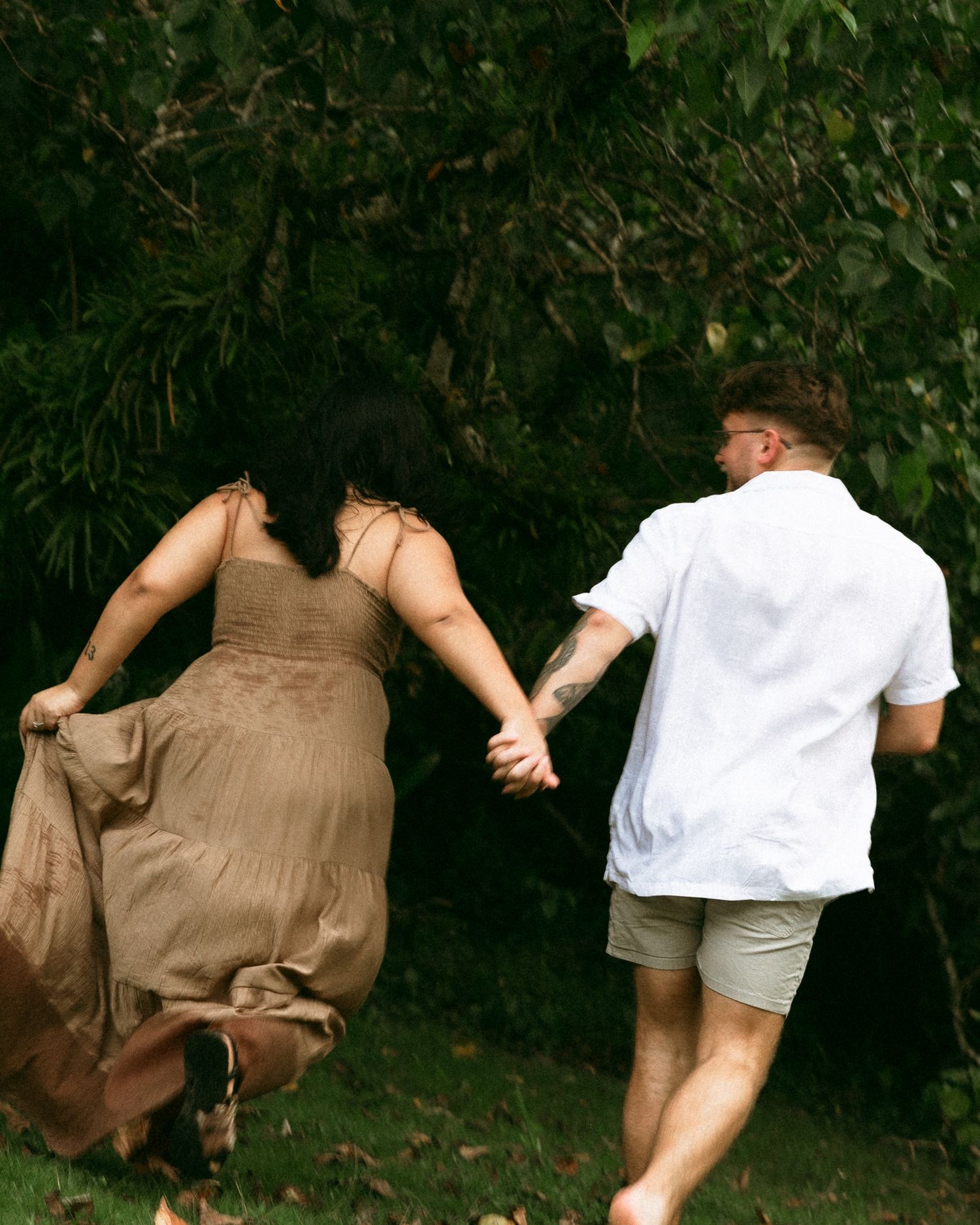 Couple holding hands walking toward lush green trees, woman in flowing tan dress, man in white shirt and gray shorts.