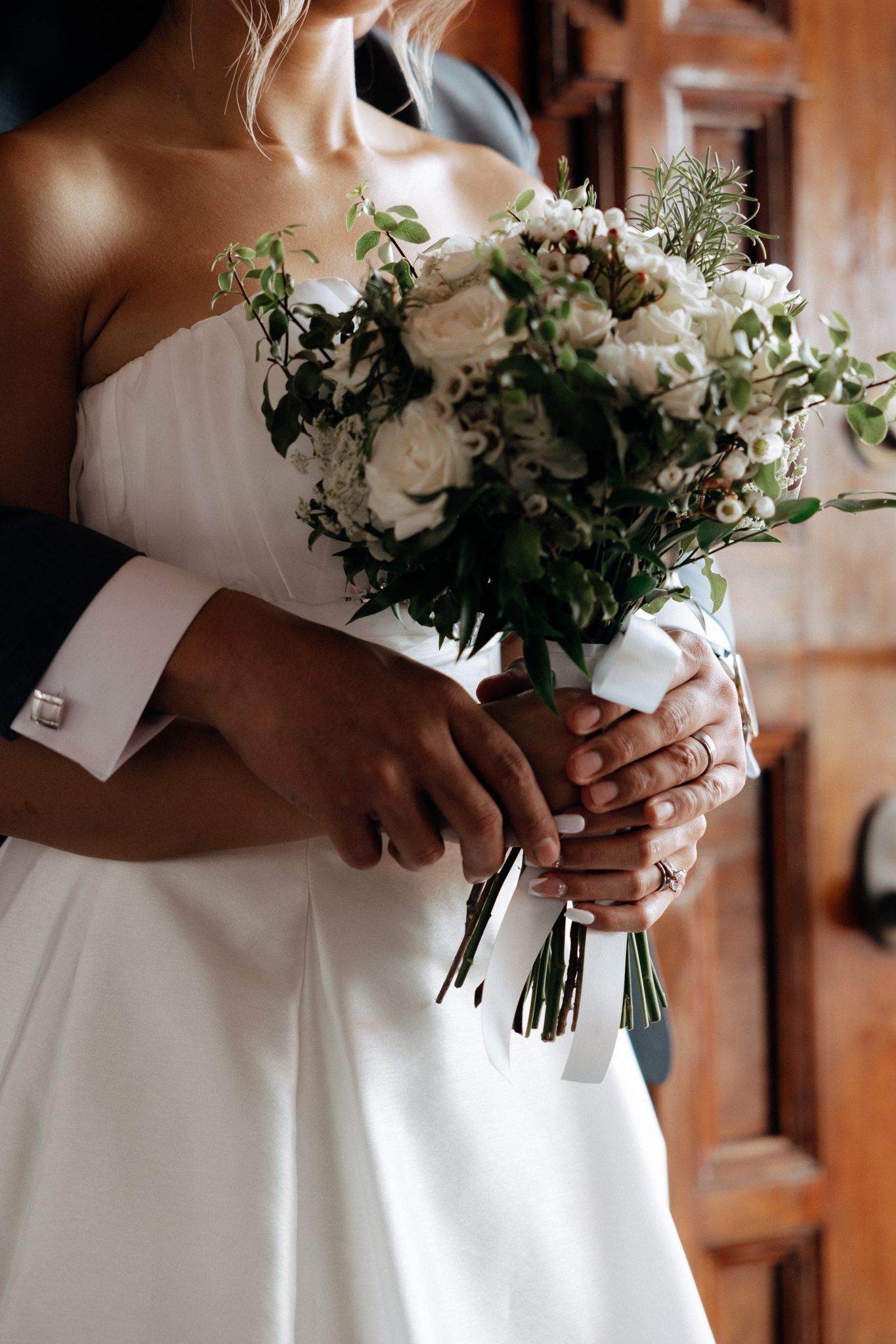Bride in white strapless gown holding elegant bouquet of white roses and greenery, embraced by groom in dark suit.