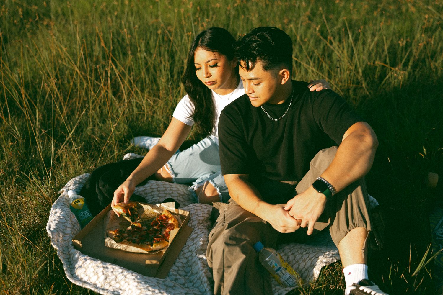 Couple enjoying a pizza picnic together in a sunny grassy field, sharing a romantic outdoor meal.