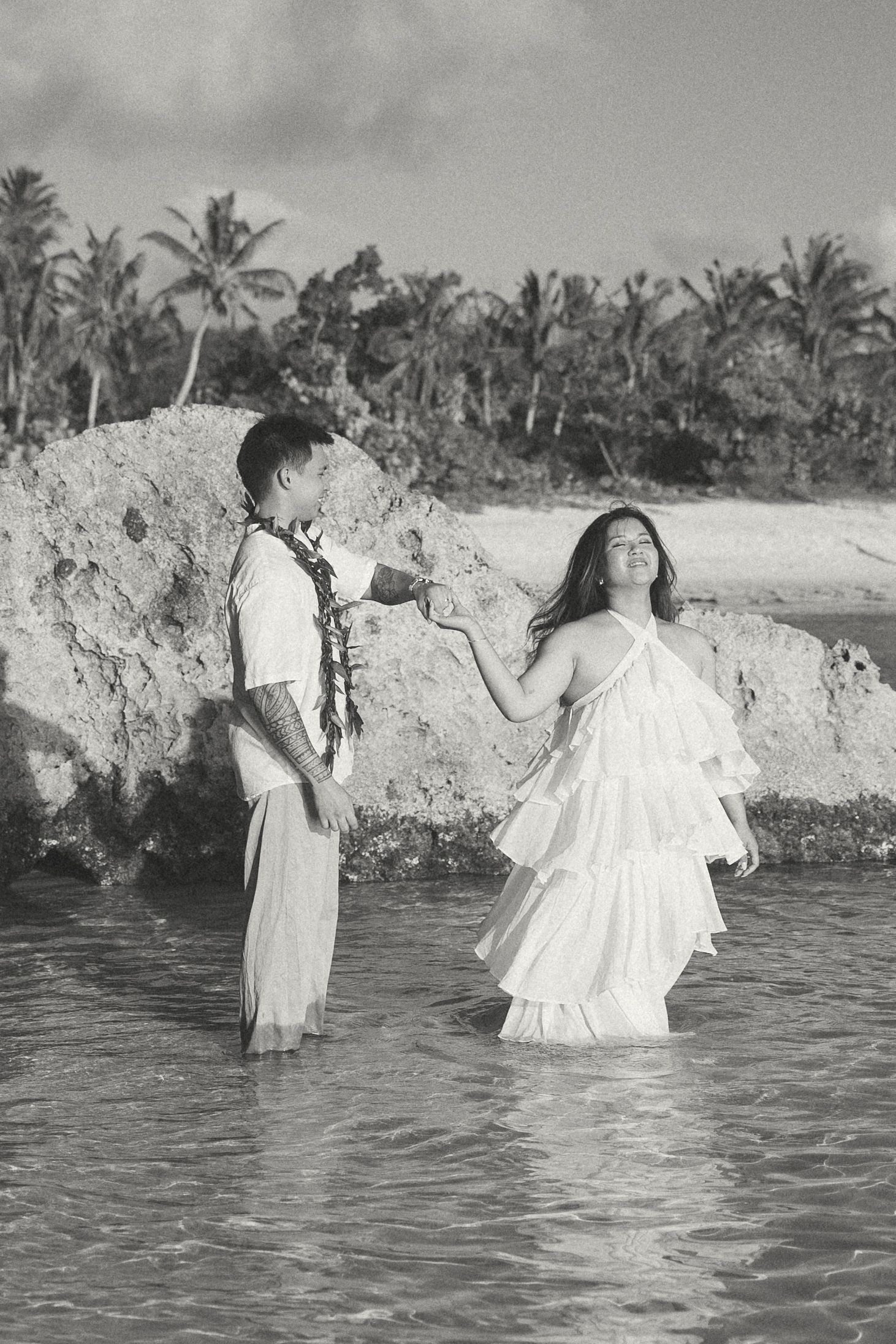 Black and white photo of a couple in white attire standing in ocean water near a tropical beach with palm trees.