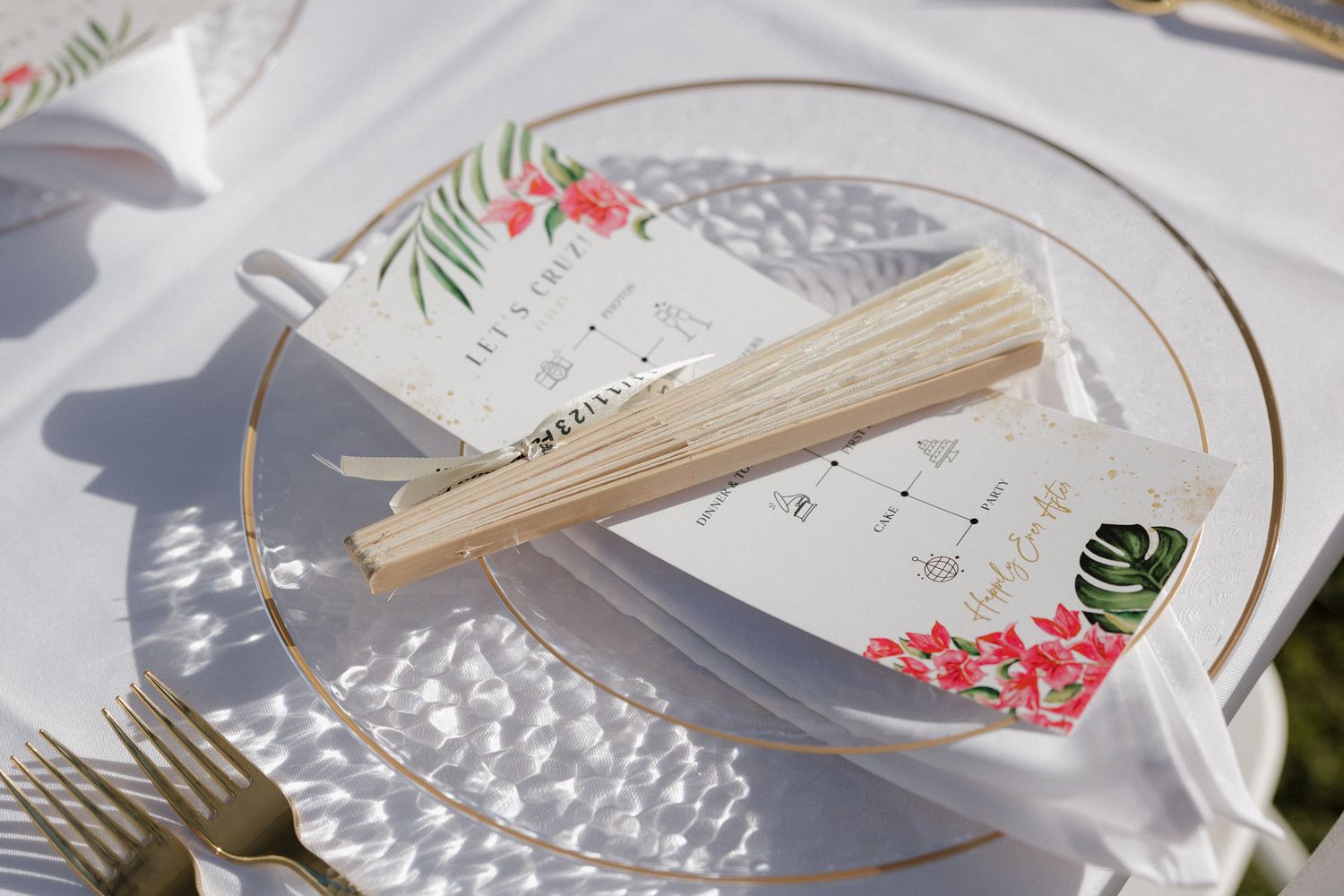 Elegant tropical wedding place setting with fan, floral menu card on gold-rimmed glass plate, white linen table.