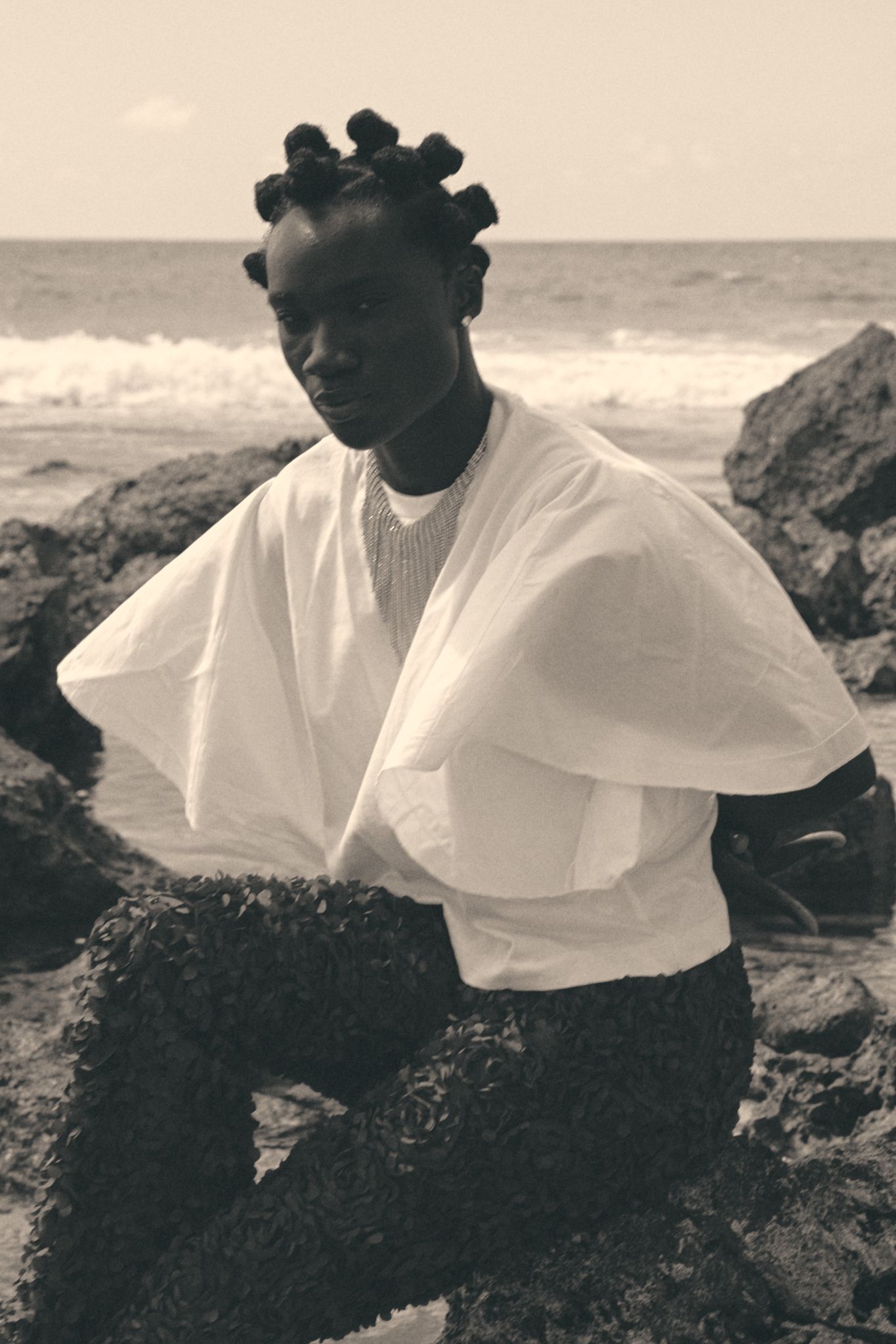 A model with bantu knots wears a white draped top and dark skirt, posing on rocky beach in sepia toned photo.