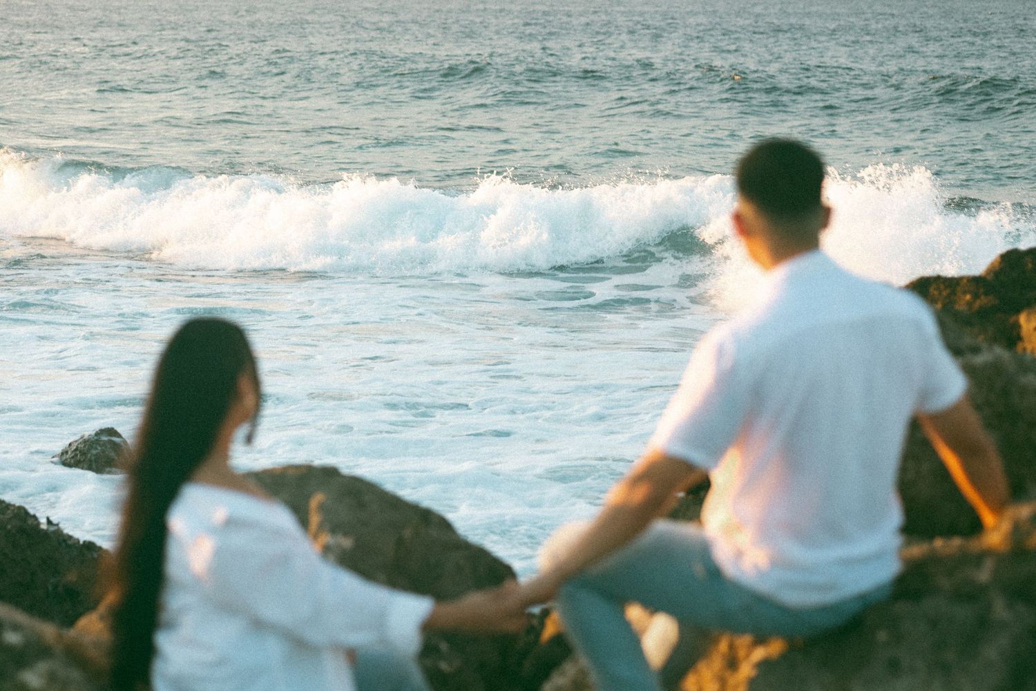 Couple sitting on rocky shore watching ocean waves at sunset, wearing white outfits in a romantic seaside setting.
