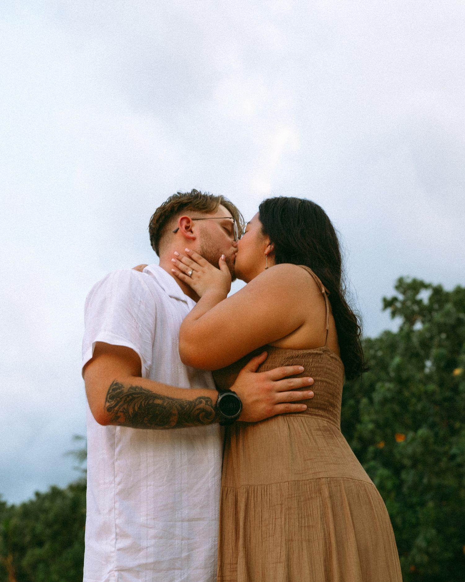 A couple shares a romantic kiss outdoors, man in white shirt with tattoos, woman in tan dress, trees in background.