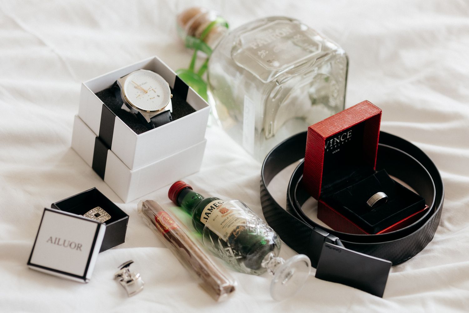 A flat lay of groom accessories including a watch, belt, cufflinks, ring box, cigar, and liquor bottles on white fabric.