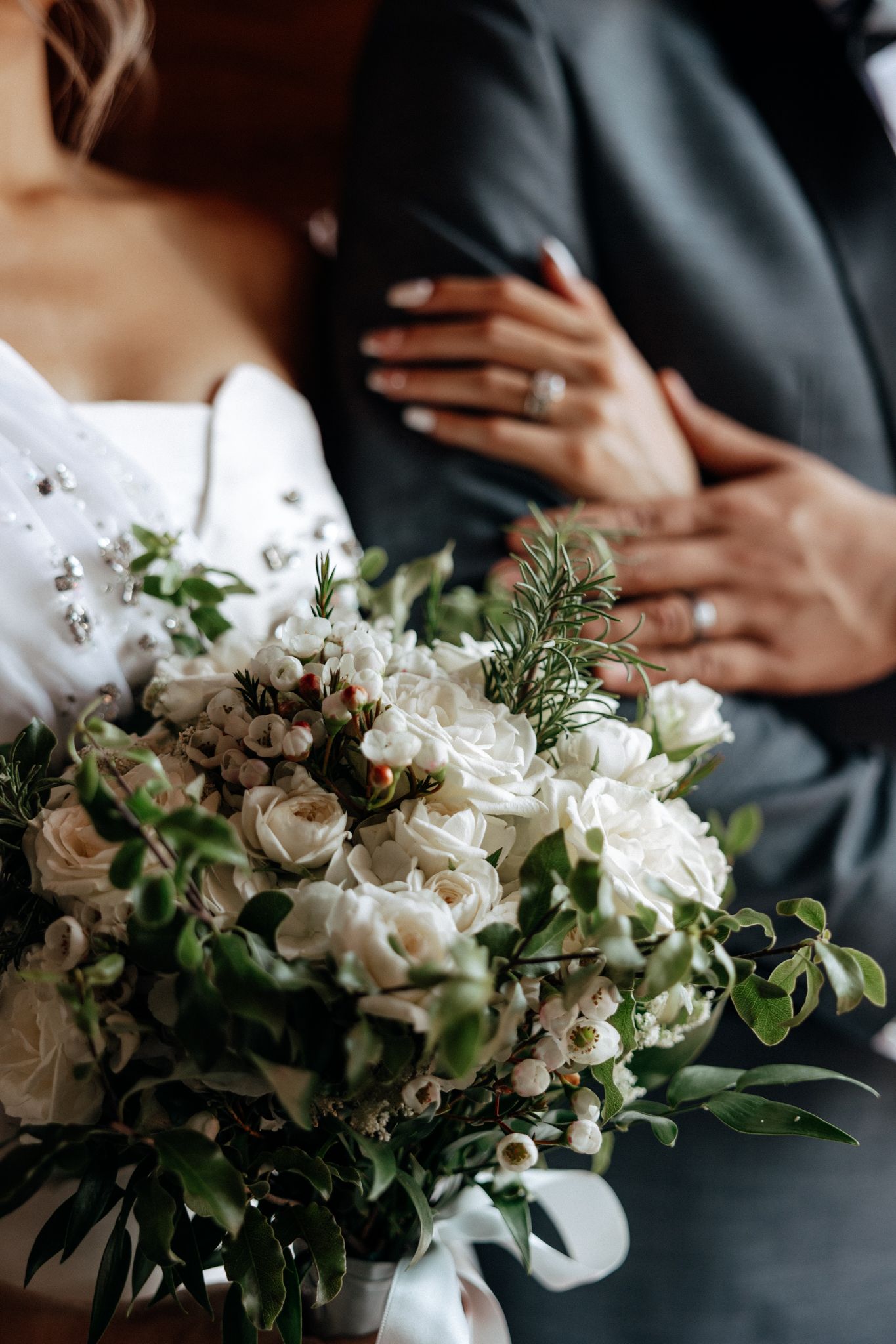 Bride holding white rose bouquet with greenery while groom embraces her, both wearing wedding rings.