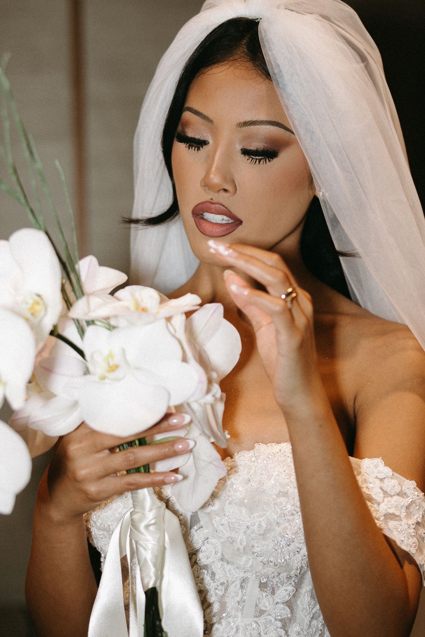 Elegant bride in white lace gown and veil holds orchid bouquet, showcasing glamorous makeup and manicured nails.