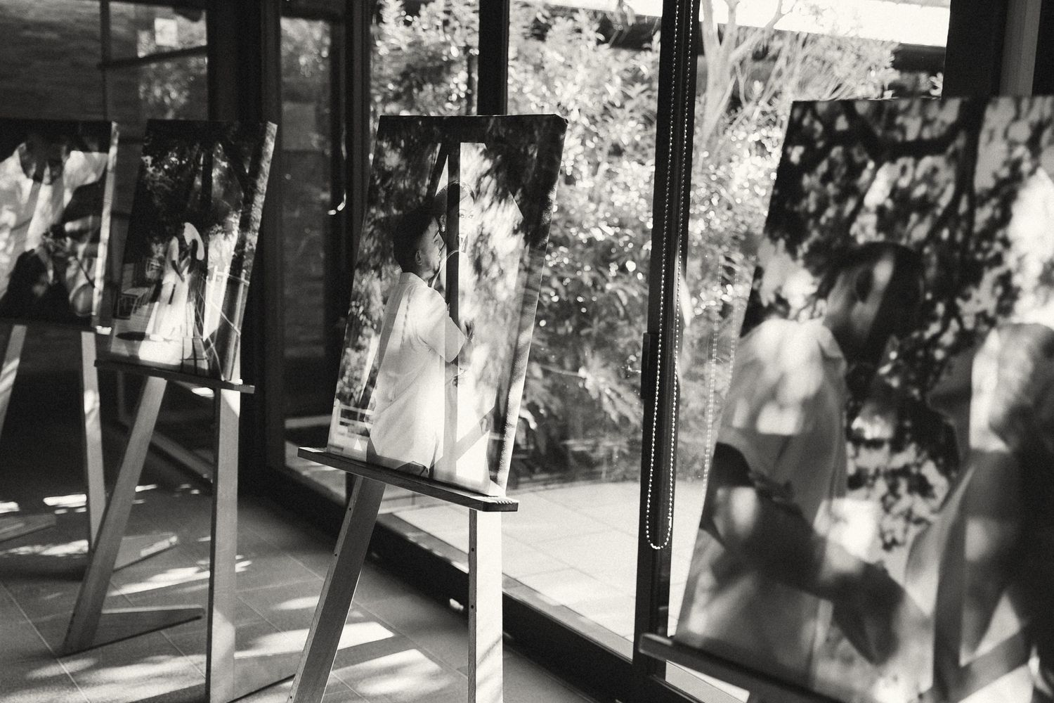Black and white photo of mirrors on easels reflecting a woman in white dress amid lush garden views through large windows.