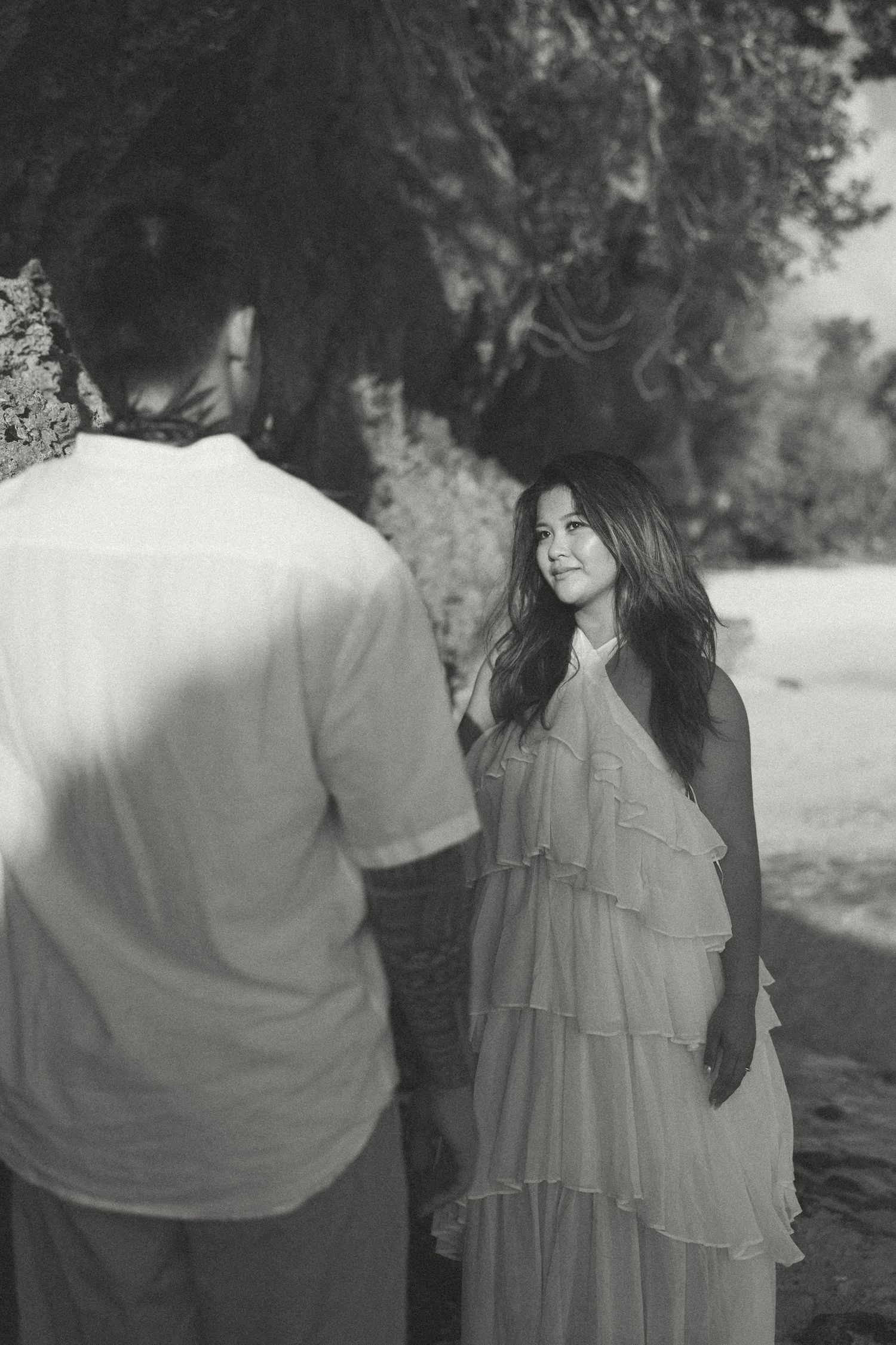 Black and white photo of a woman in a flowing tiered dress smiling at a man outdoors near a tree.