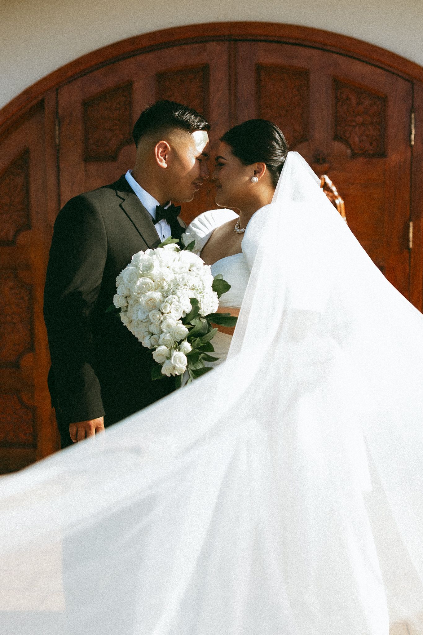 Bride and groom share a romantic moment, her flowing veil billowing, holding white bouquet before ornate wooden doors.