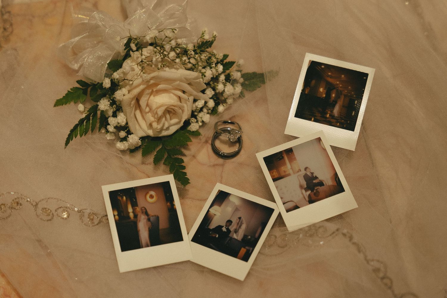 Wedding flat lay featuring Polaroid photos, white rose boutonniere, and diamond rings on lace fabric.