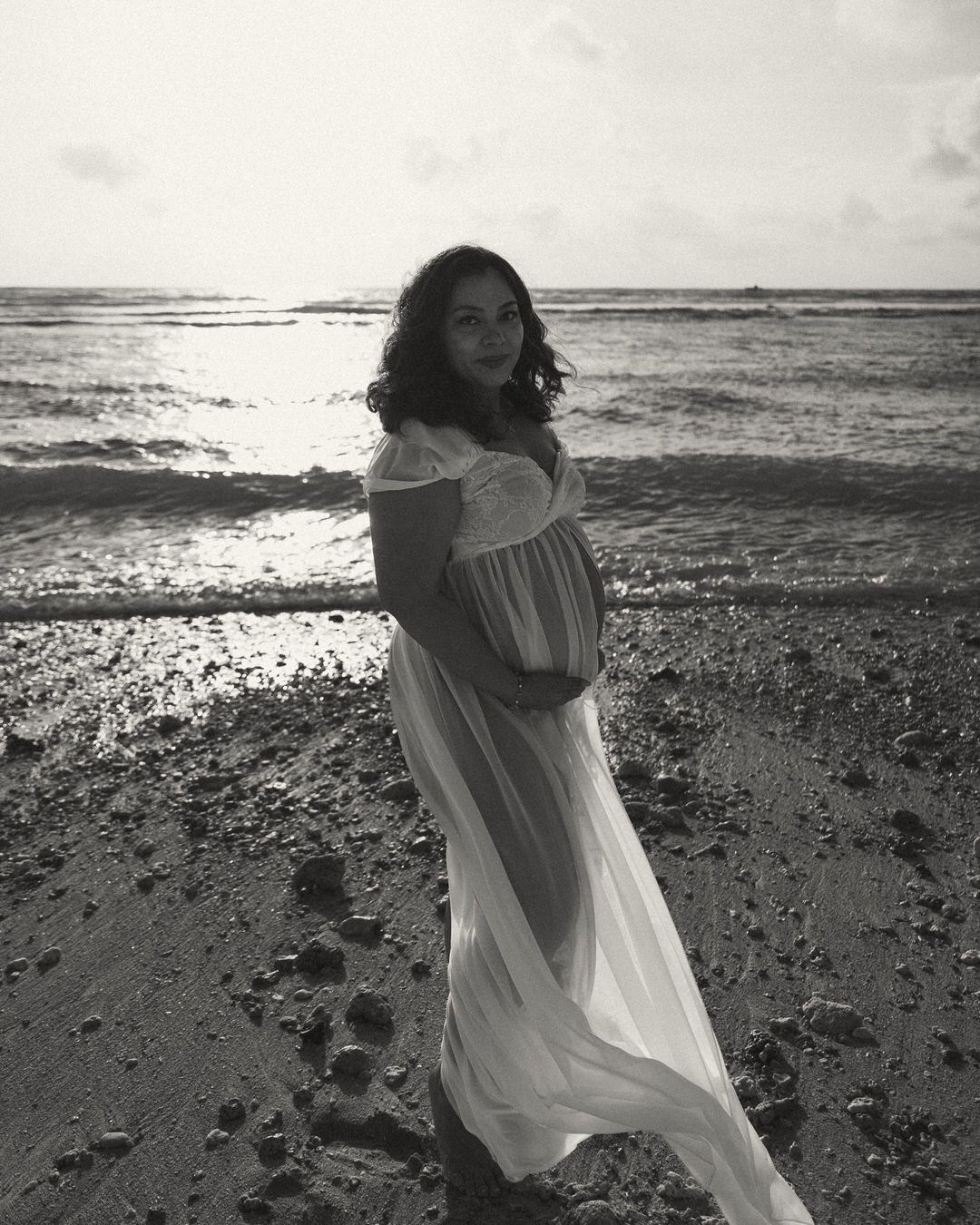 Pregnant woman in white flowing dress standing on beach shore in black and white maternity photo.