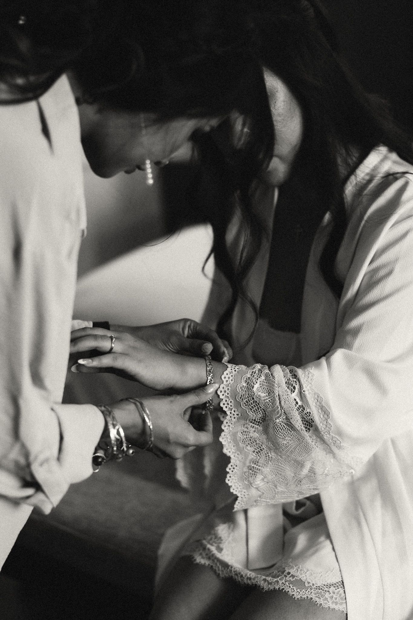 Black and white photo of woman in lace robe examining jewelry during bridal preparation.