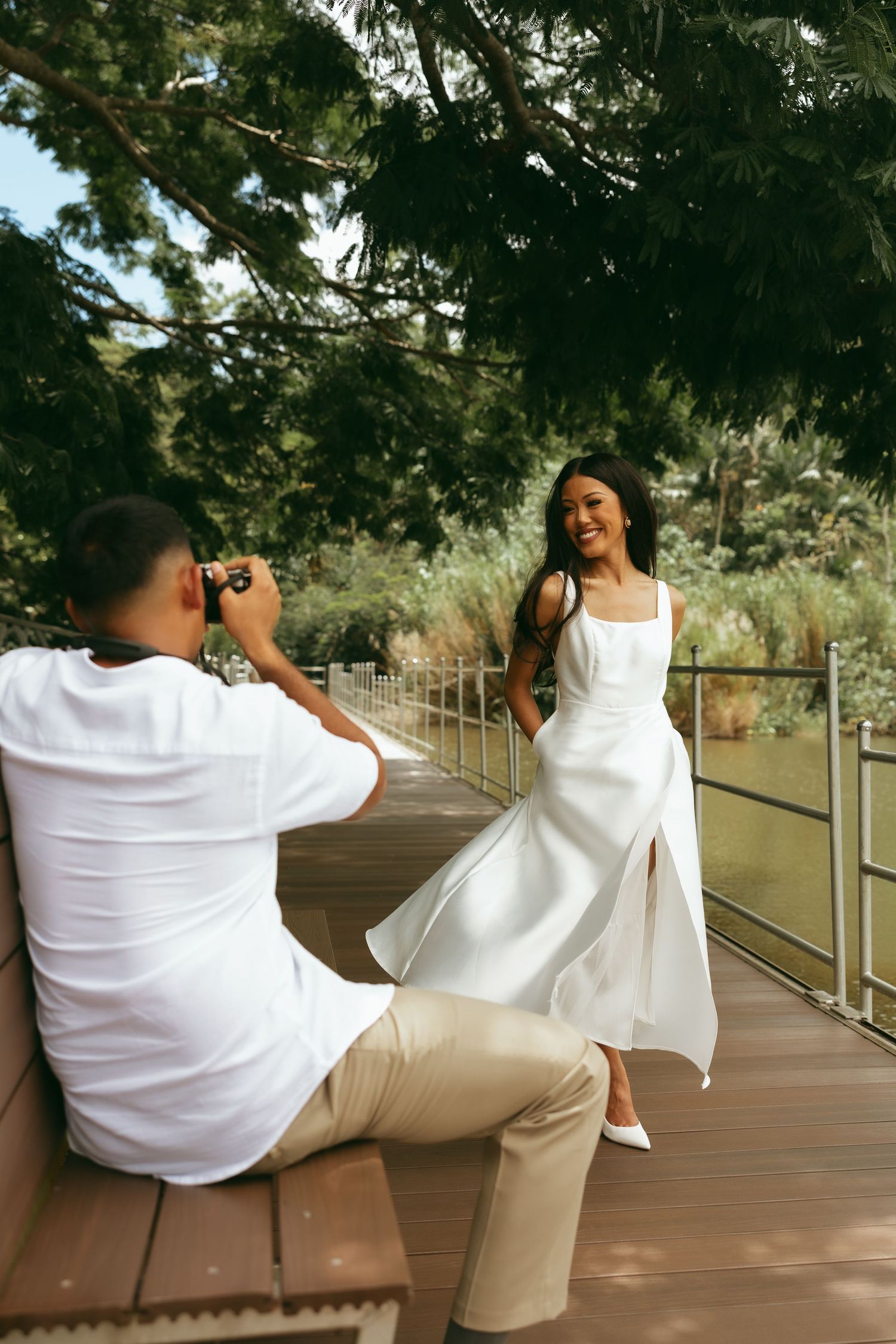 Man photographs smiling woman in white dress on wooden deck surrounded by lush green trees.