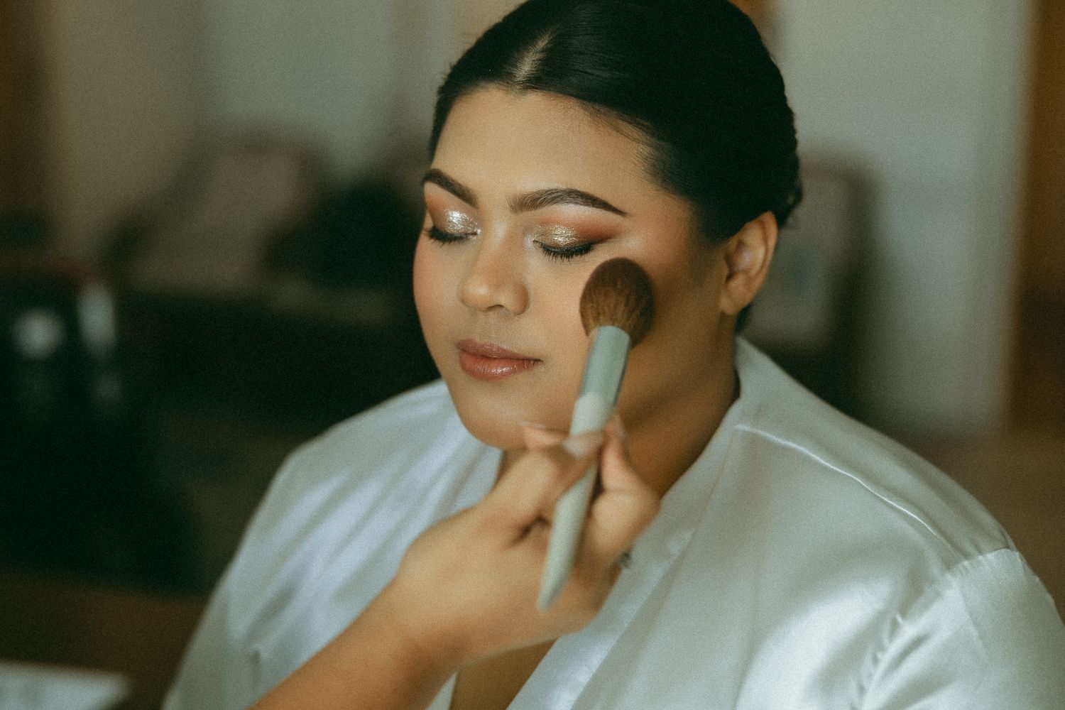 A bride getting blush applied with a makeup brush during bridal preparation in a white robe.