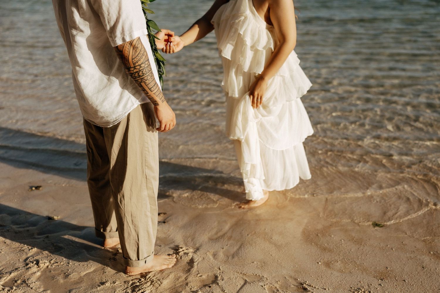 Couple holding hands on sandy beach, bride in white ruffled dress, groom in khakis with tattoos, ocean waves background.