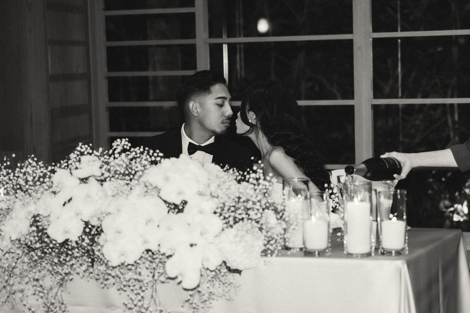Black and white wedding photo of couple sharing intimate moment at floral head table with candles.