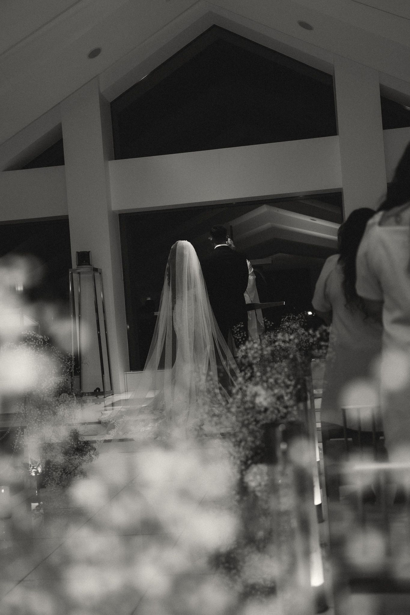Black and white photo of a bride in a veil walking down the aisle at an elegant wedding ceremony.
