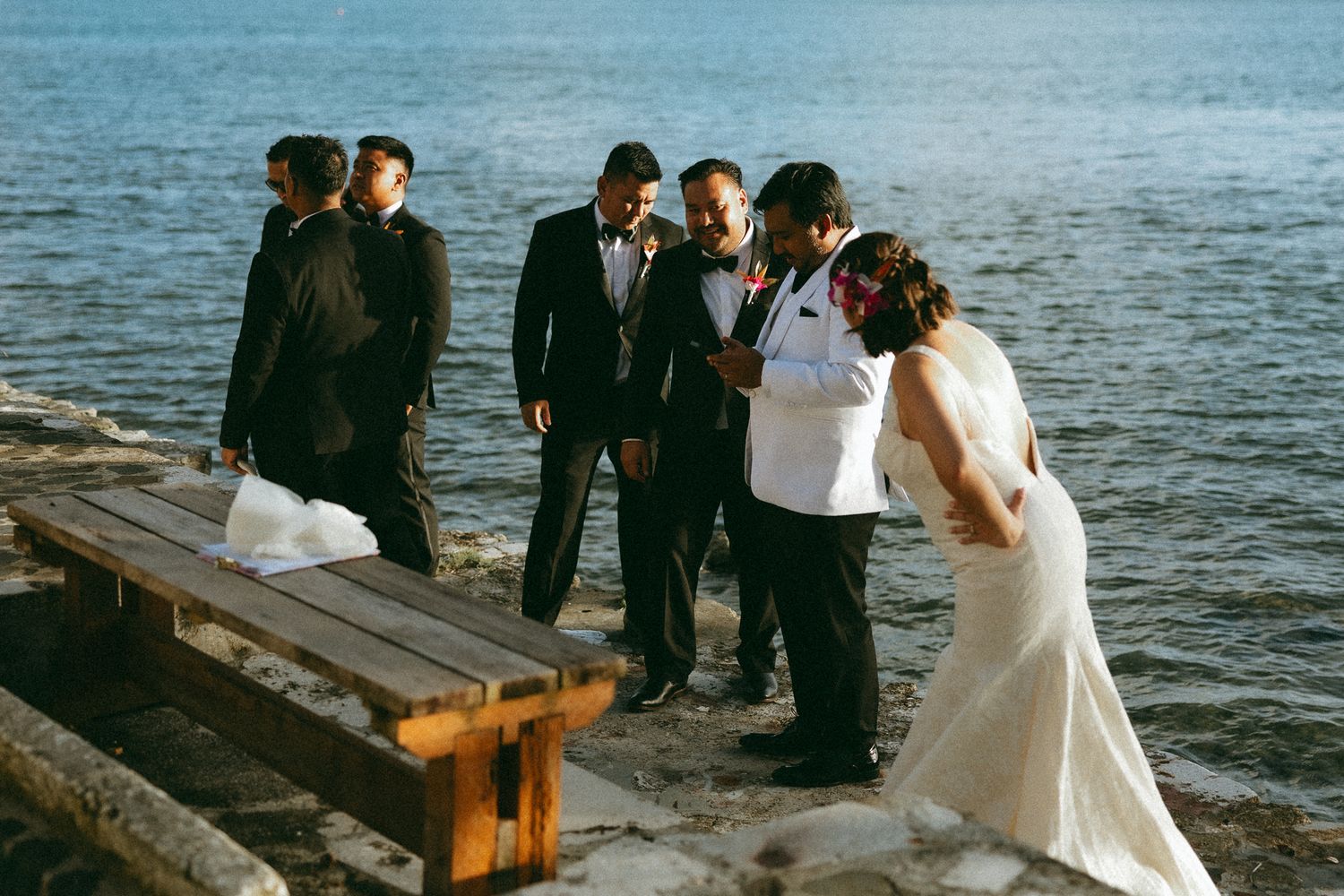 Bride and groom share a kiss near waterfront as groomsmen in tuxedos stand nearby on stone dock.