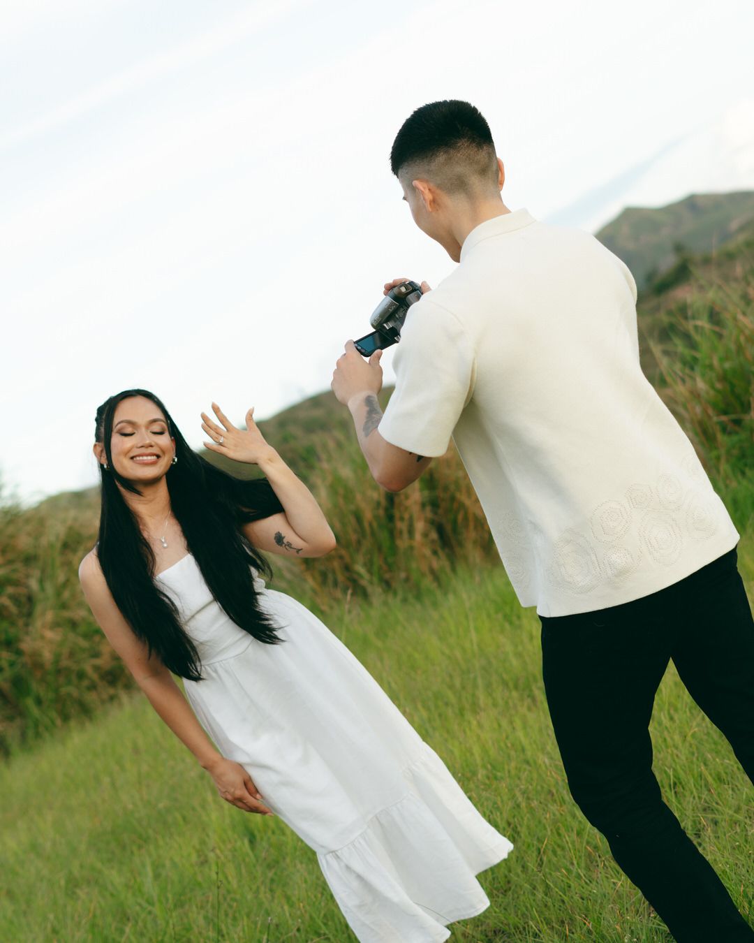 A man photographs a smiling woman in a white dress on a green hillside during an outdoor photoshoot.