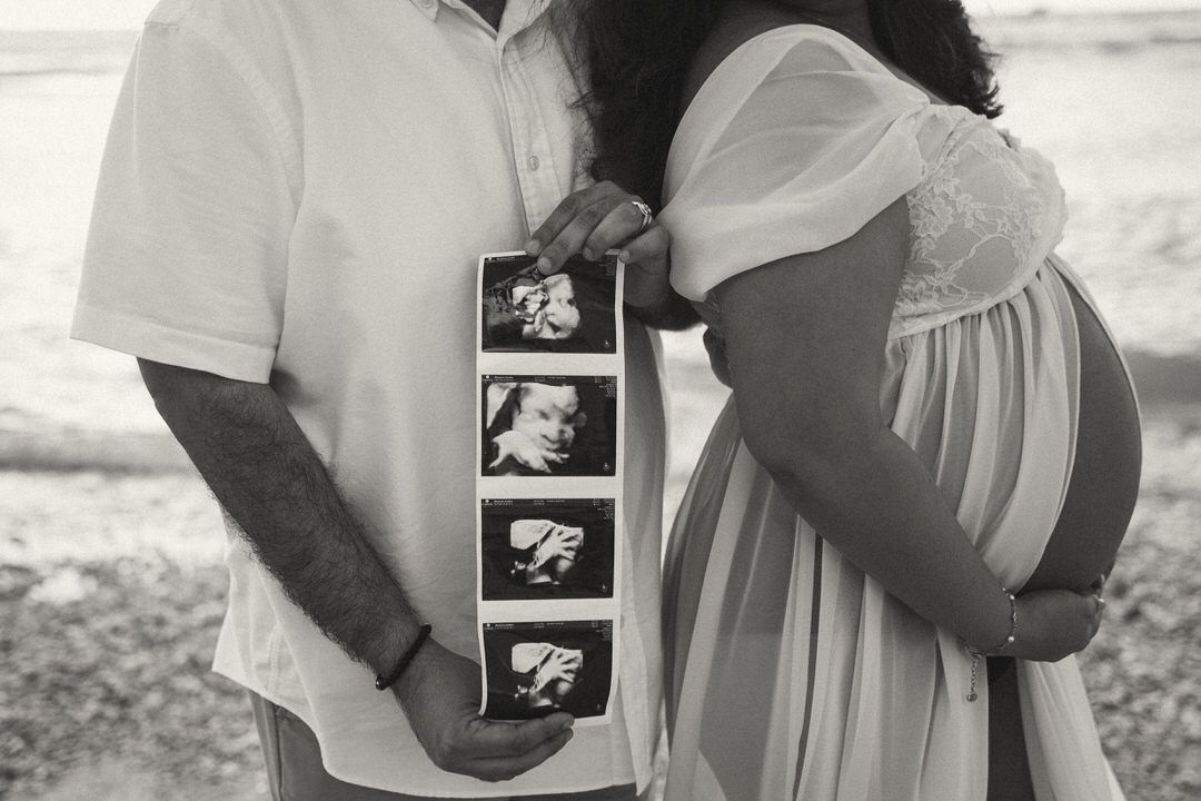 Pregnant couple holding ultrasound photo strip outdoors in black and white maternity portrait.
