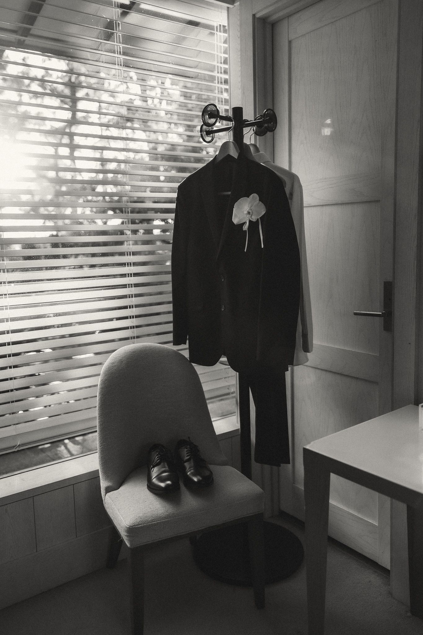 Black and white photo of a grooms suit on a hanger with boutonniere and dress shoes on a chair.