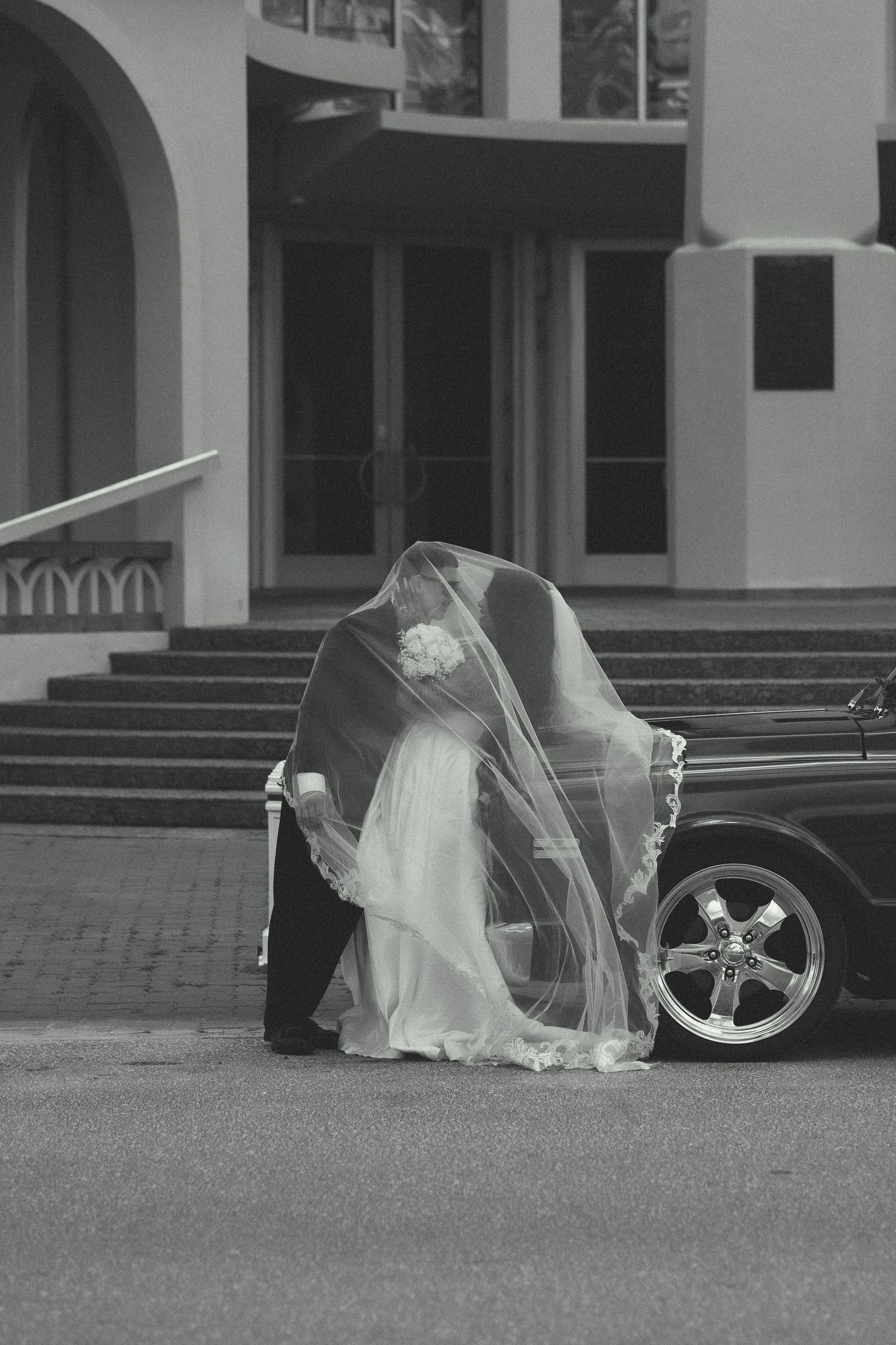 Black and white photo of bride and groom kissing beside a vintage car outside a church on wedding day.