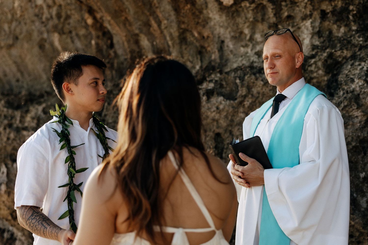 Outdoor beach wedding ceremony with groom in white shirt and lei, bride, and officiant in teal sash holding a book.