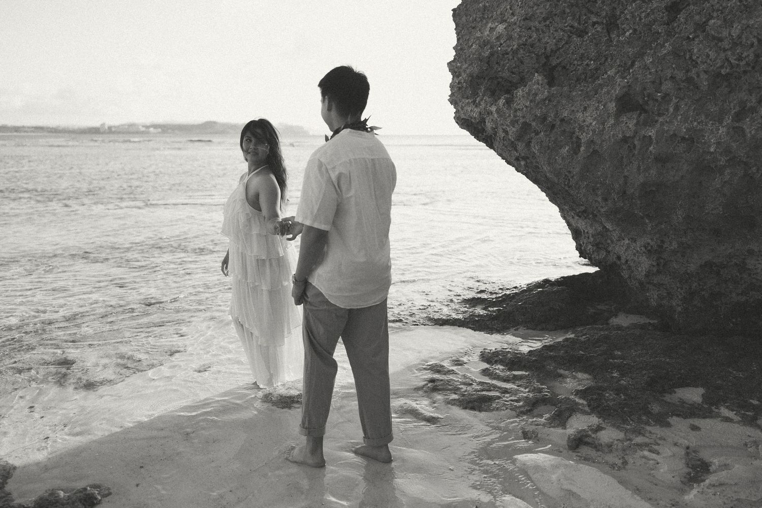 Black and white photo of a couple holding hands on a rocky beach shore in white attire.
