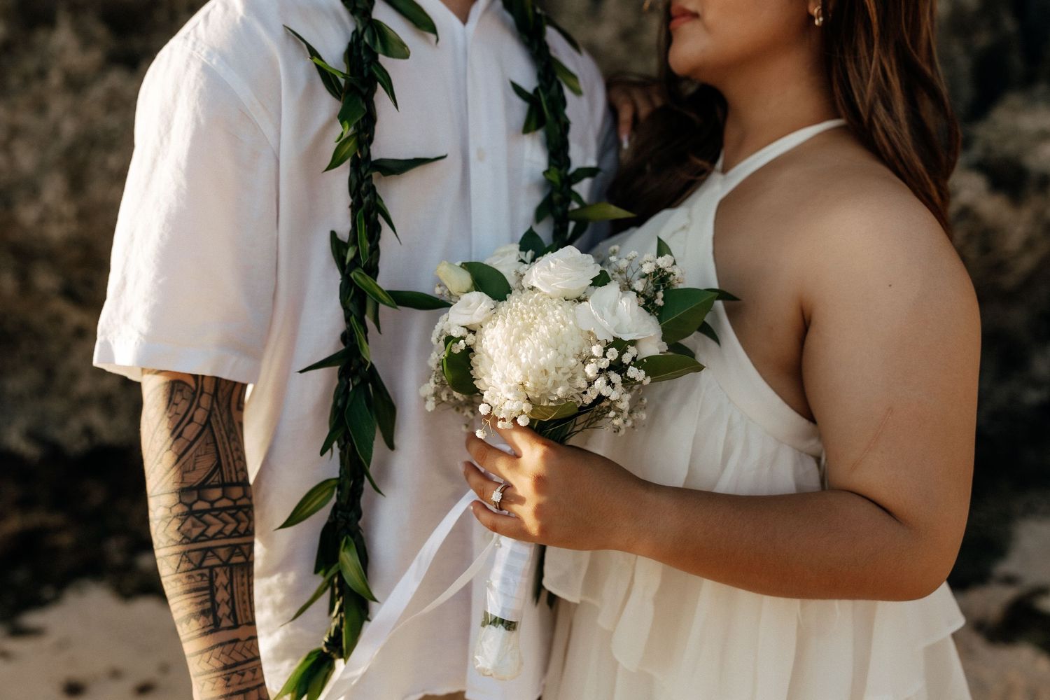 Couple in wedding attire on beach, groom with floral lei and tattoos, bride holding white bouquet in flowing dress.