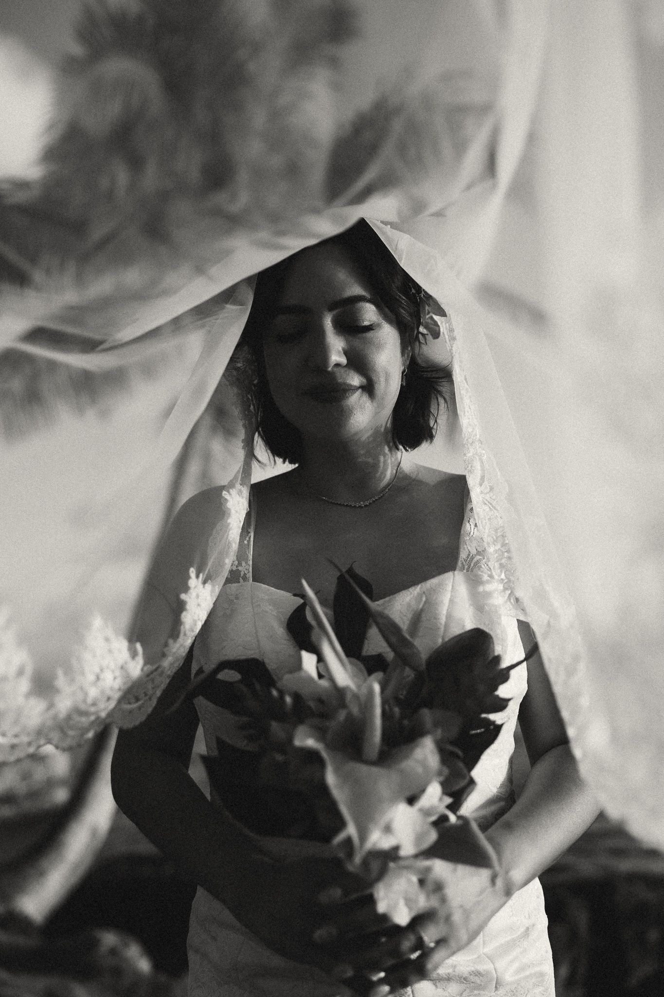 Black and white photo of a bride holding a bouquet, eyes closed, with a flowing veil draped over her head.