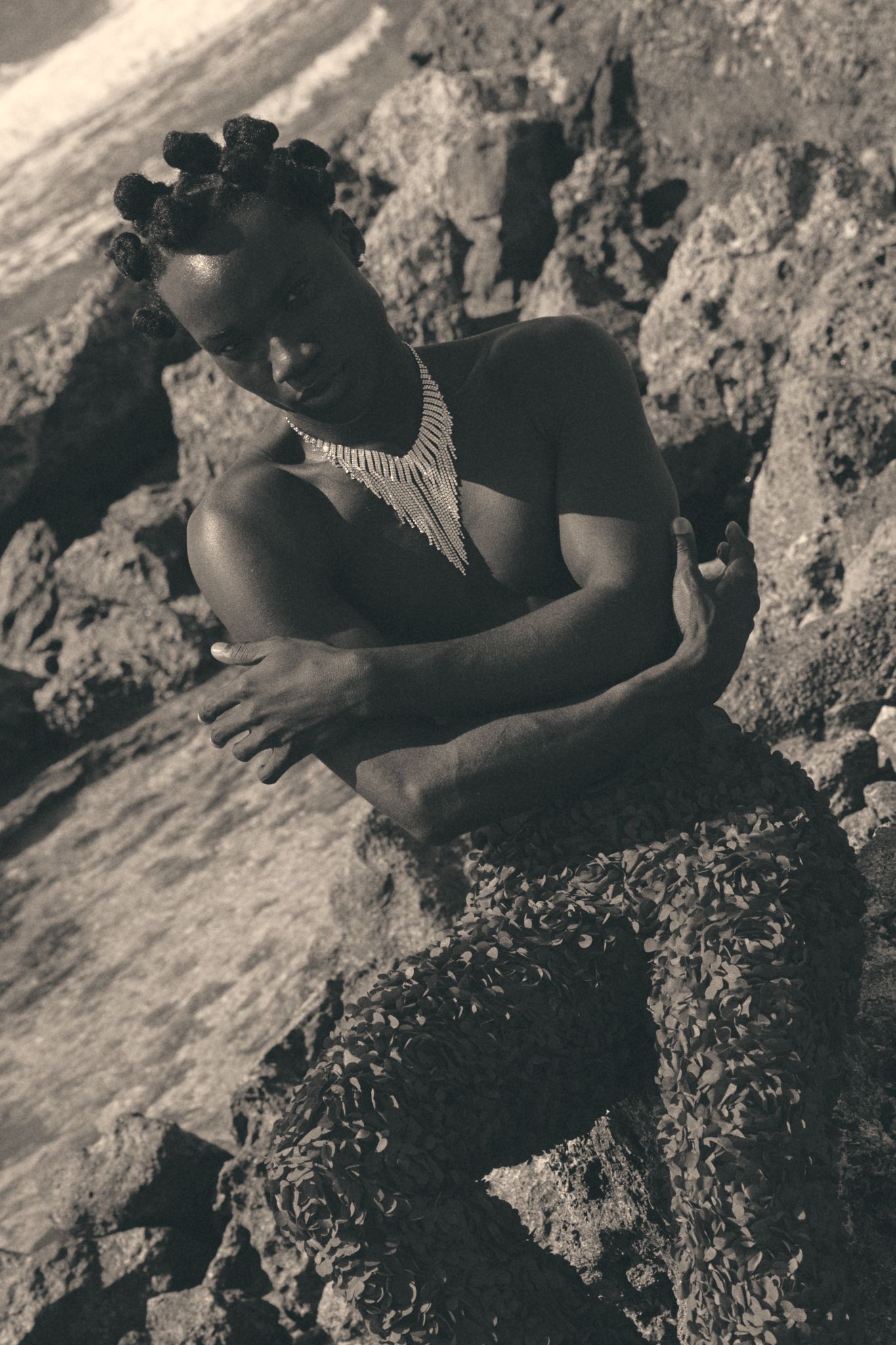 Black and white photo of a woman in a statement necklace posing gracefully on rocky coastal terrain.