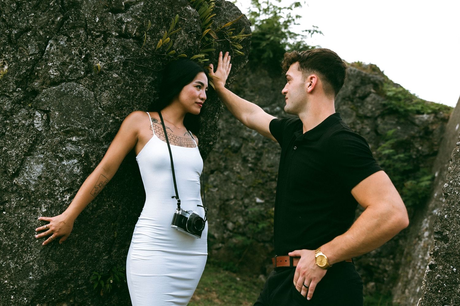 A man and woman share an intimate moment against a rocky wall, she wears a white dress with a camera.
