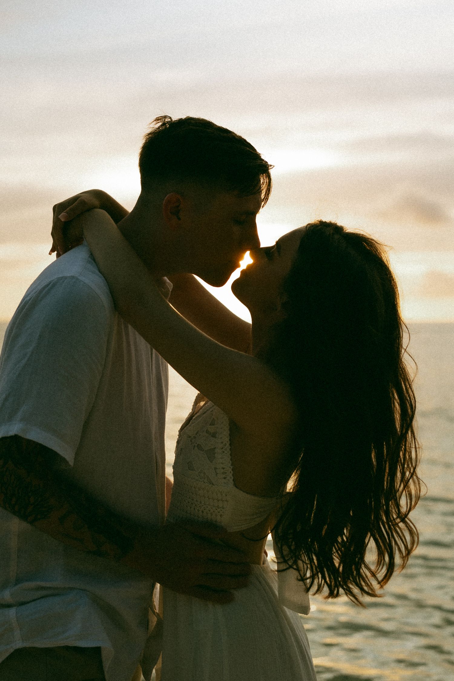 Romantic couple sharing a kiss at sunset by the ocean, silhouetted against a golden sky.