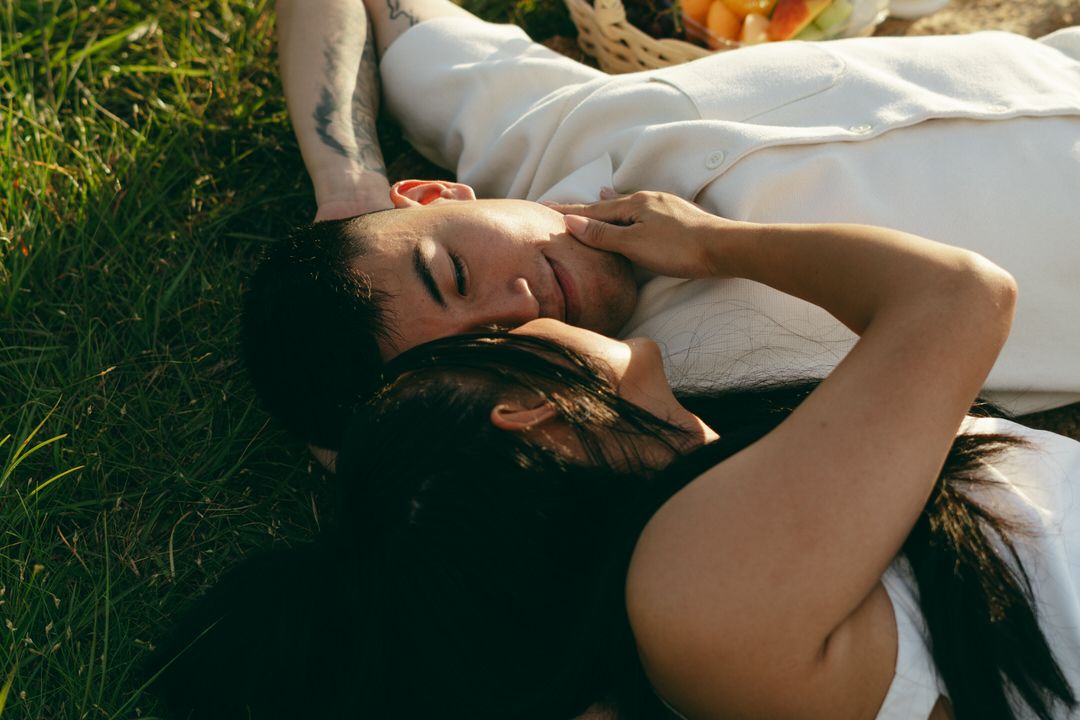 Couple lying on green grass in warm sunlight, sharing a tender romantic moment outdoors.