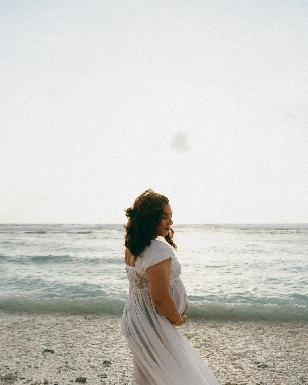 Pregnant woman in white maternity dress standing on sandy beach with calm ocean in background.