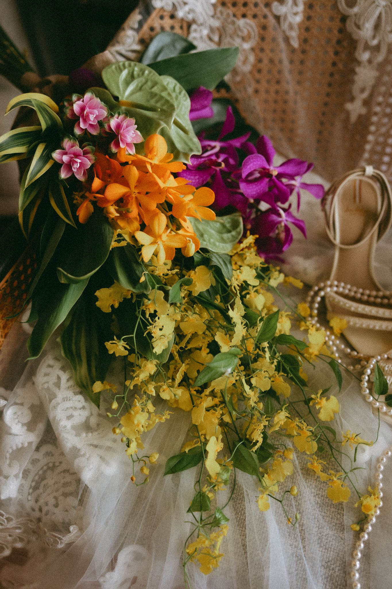 Colorful cascading wedding bouquet with yellow, orange, pink, and purple orchids on white lace dress.