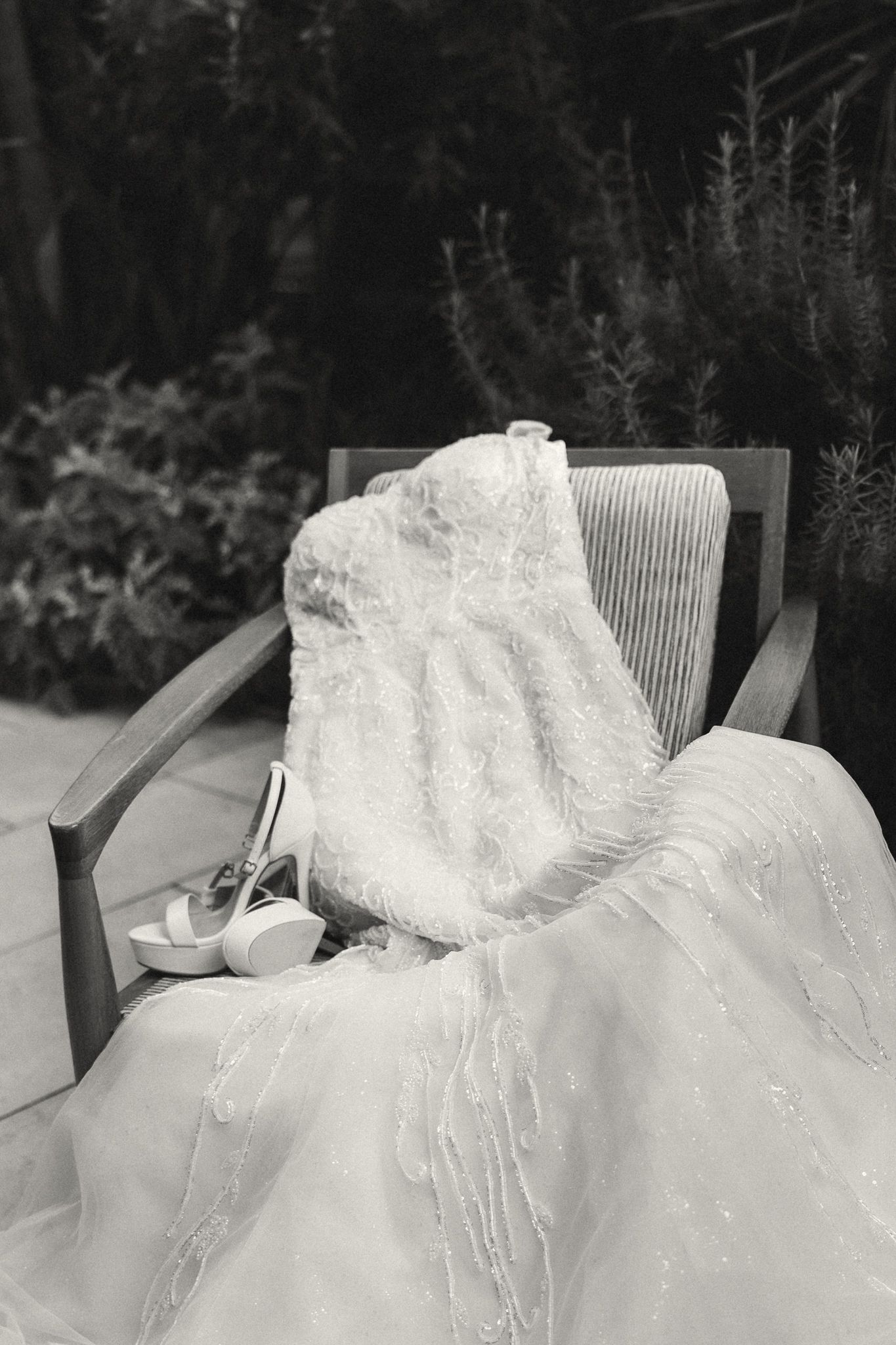 Black and white image of a lace wedding dress draped over a wooden chair with bridal heels.