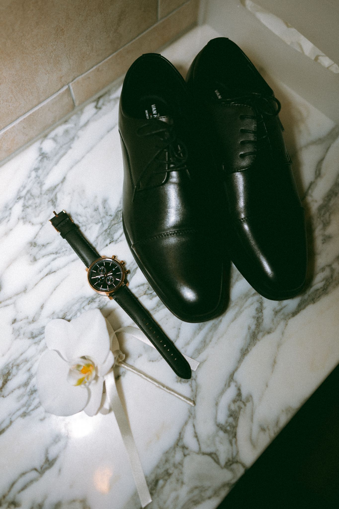 Black leather dress shoes and watch with white orchid boutonniere on marble surface.