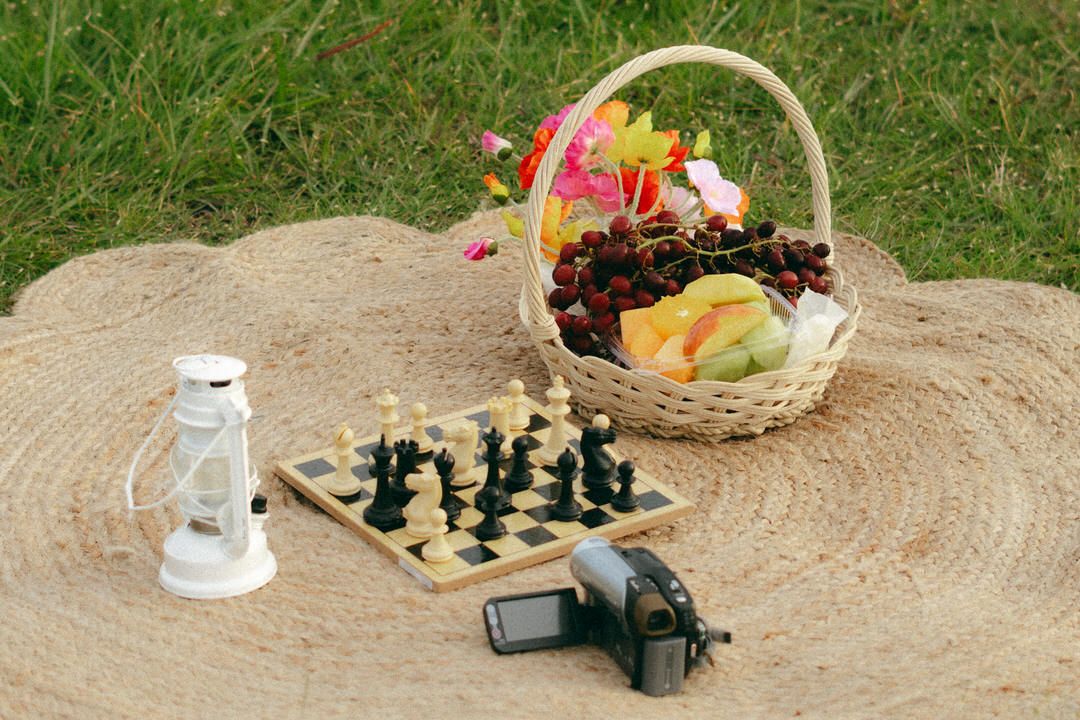 Picnic setup on a woven mat with chess board, lantern, camera, and fruit basket on grass.
